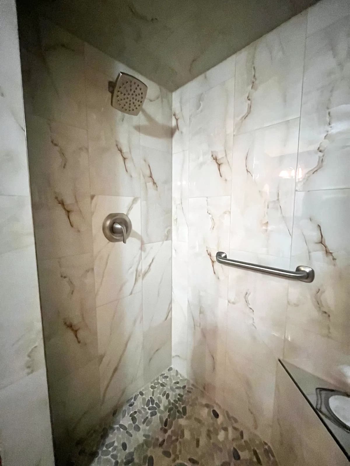 Rockwall, TX shower with marble-look large-format wall tile, a pebble-stone floor, a tiled ledge and a grab bar